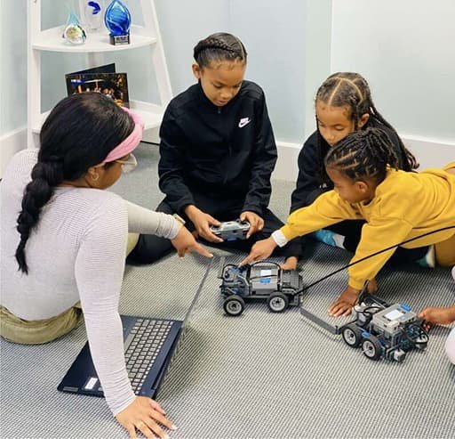 Children participating in a robotics activity at Connectech Bermuda, using laptops and remote controls to guide small wheeled robots on the floor.